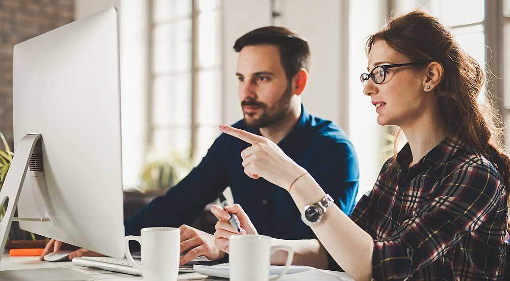 Homem e mulher em um escritório, olhando para a tela de um computador e com alguns papéis em mãos para fazer a administração de recursos humanos com a Sem Parar Empresas