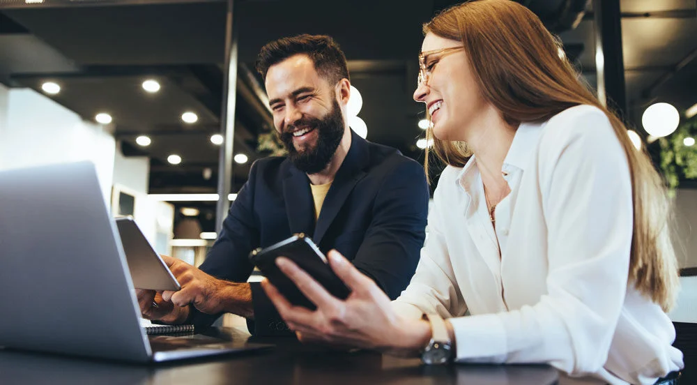 emissao_recibo_de_vale_transporte_sem_parar_empresasHomem e mulher sorrindo em um escritório, fazendo a emissão do recibo de vale-transporte Sem Parar Empresas em notebooks e tablets