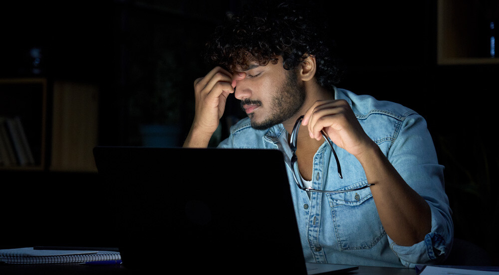 Sem Parar Empresas: homem em sala escura, na frente de um notebook, com a mão no rosto e aspecto cansado, indicando que está passando pela tendência quiet quitting