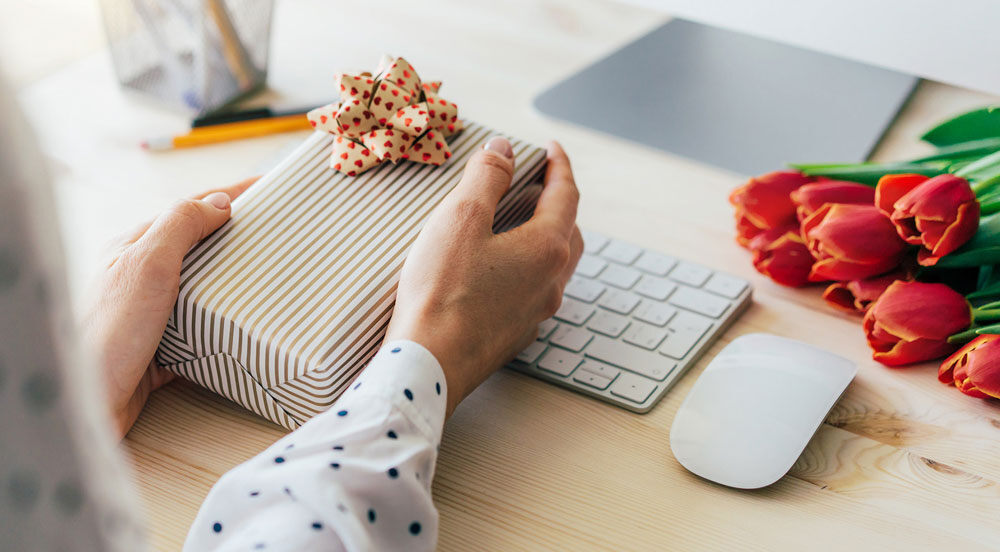 Sem parar Empresas: mãos segurando presente em cima da mesa de trabalho com um computador e buquê de flores, representando uma mensagem dia das mães empresa