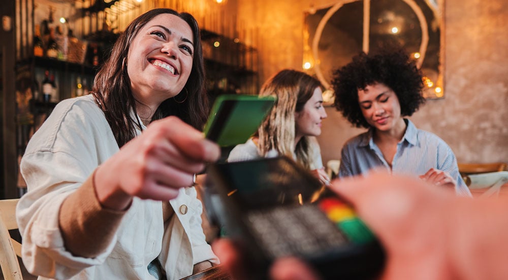 Mulher sorrindo pagando com um cartão multibeneficios e duas outras mulheres ao fundo.