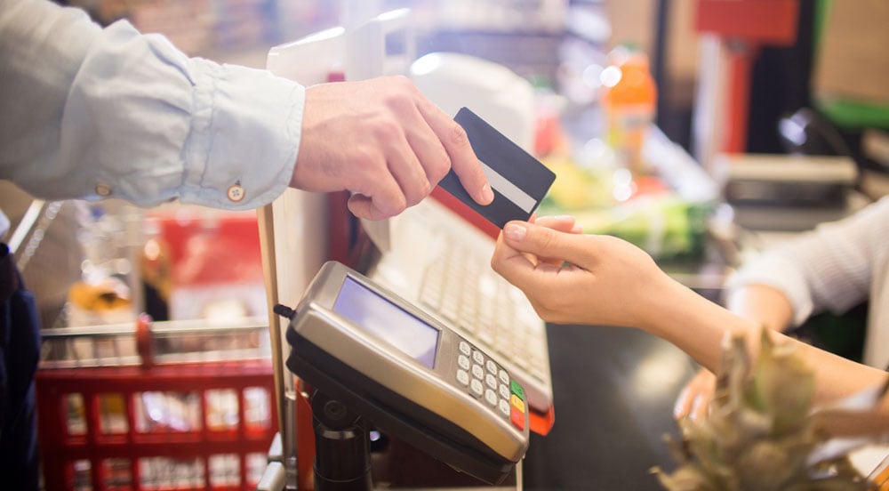 Uma mão passando um cartão de pagamento para outra pessoa, que é um caixa de supermercado
