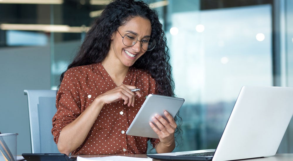 Mulher sorrindo em escritório em frente a um computador, segurando um tablet para verificar suas ferramentas de gestão Sem Parar Empresas