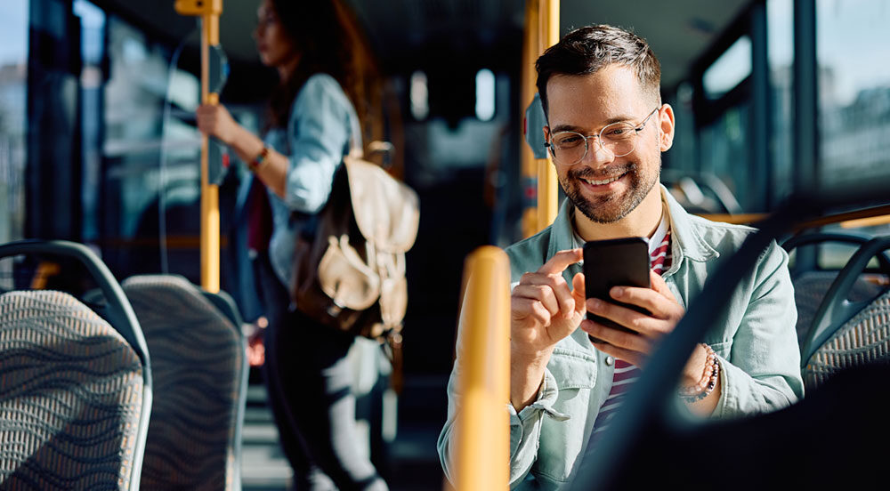 Homem sentado em banco do ônibus, mexendo no celular e sorrindo, pois utiliza o vale-transporte CLT da Sem Parar Empresas