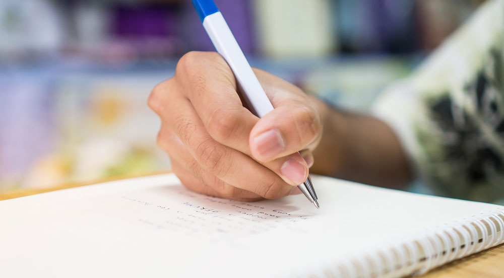 Mão escrevendo à caneta em um caderno, representando estudante que utiliza auxílio-educação fornecido pelo Cartão Multi da Sem Parar Empresas