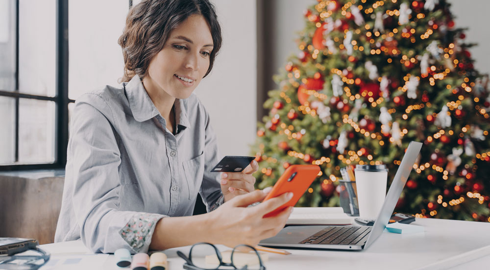 Sem Parar Empresas: mulher trabalhando em casa em frente a uma árvore de Natal, com celular e um cartão na mão, onde recebe seu kit de Natal para empresas como benefício flexível