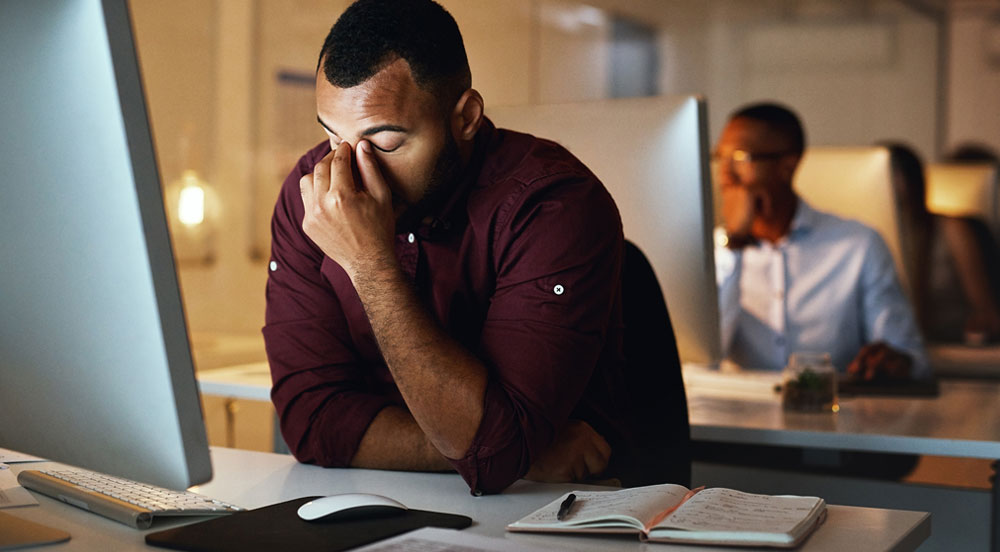 Sem Parar Empresas: homem em escritório, em frente ao computador, com aspecto cansado e esfregando os olhos, indicando que pode estar em uma crise de burnout