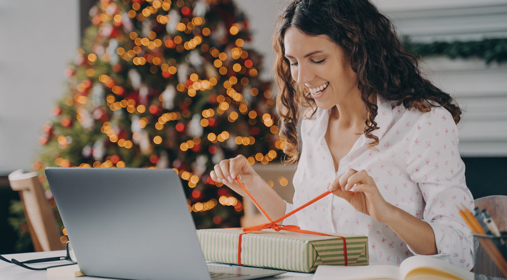 Sem Parar Empresas: mulher membro de equipe remota abrindo um presente na frente de um computador e com uma árvore de Natal ao fundo