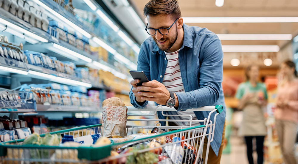 Sem Parar Empresas: homem em supermercado debruçado sobre o carrinho enquanto consulta no celular se pode utilizar VA e VR em suas compras