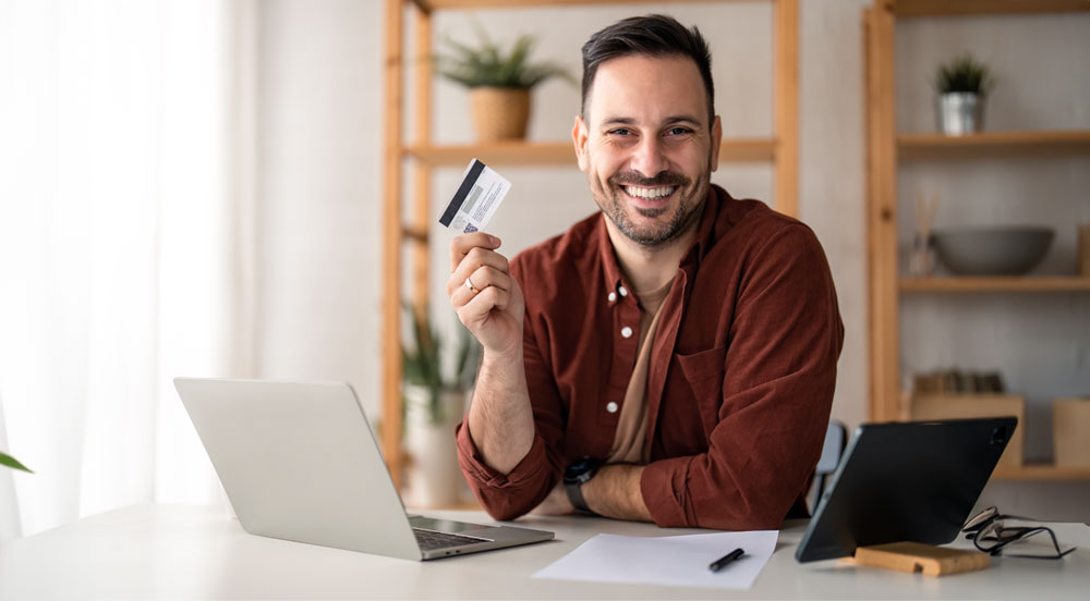 Homem sorrindo sentado em uma bancada com um computador e segurando o cartão Multi Sem Parar Empresas na mão