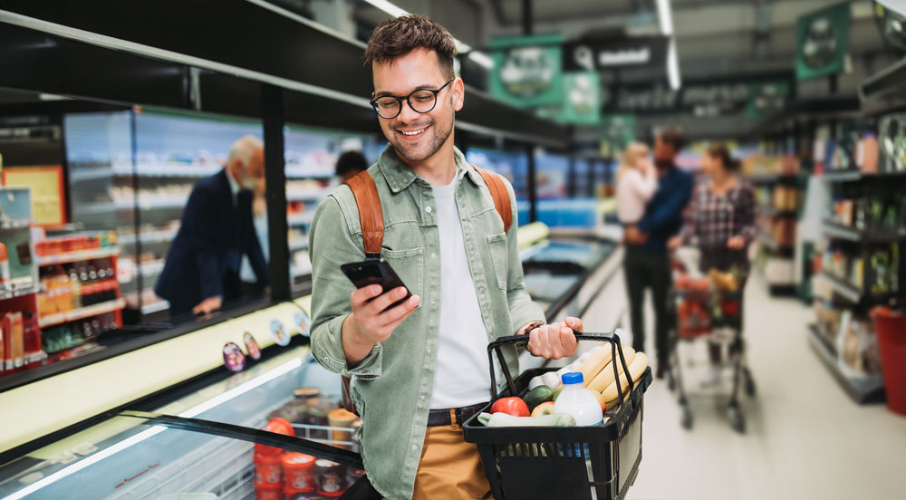 Homem em supermercado com um celular em uma das mãos e na outra uma cesta com alimentos comprados com vale-alimentação Sem Parar Empresas