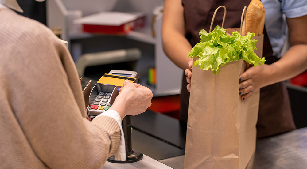 pessoa fazendo pagamento via cartão em caixa de supermercado, onde está passando uma sacola de papel com um alface e uma baguete