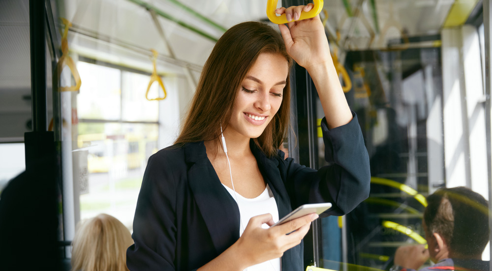 Mulher sorrindo em pé em um ônibus, segurando o celular e com fones de ouvido