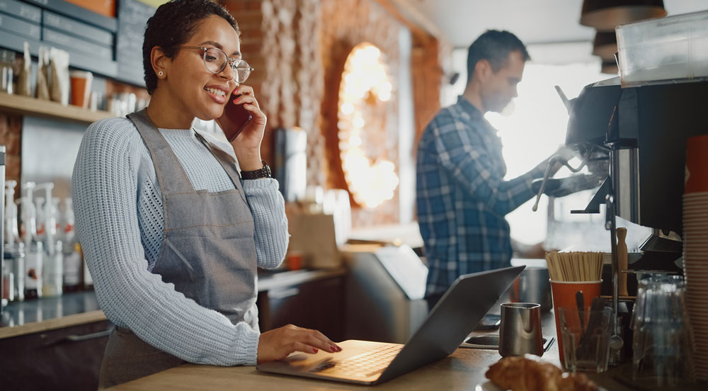 Sem Parar Empresas: mulher falando no telefone e mexendo no notebook para verificar os benefícios extras para pequenas empresas que estão disponíveis