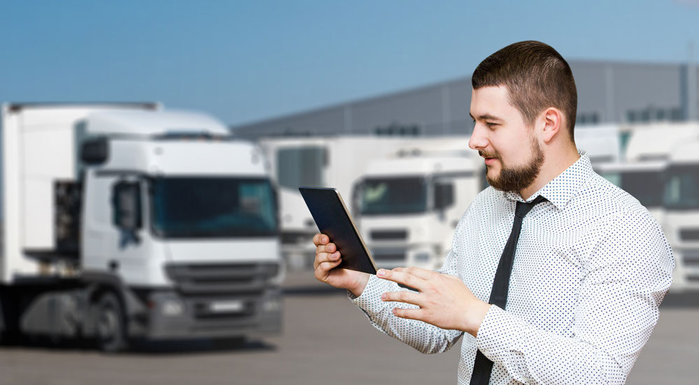 Sem Parar Empresas: um homem de gravata em um pátio com caminhões estacionados olhando a tela de um tablet para fazer gestão de transporte