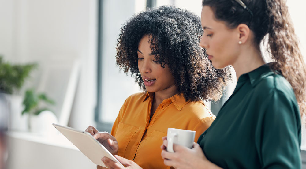 Sem Parar Empresas: duas mulheres conversando no escritório, uma com um café e outra com um tablet para fazer a gestão de pedágio da frota