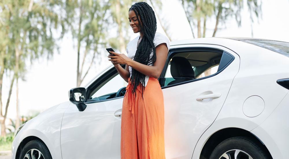 Mulher em frente a um carro branco olhando seu celular para aprender como imprimir o CRLV Digital de Pessoa Jurídica com a Sem Parar Empresas