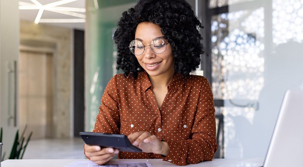 Sem Parar Empresas: mulher em frente do computador com calculadora, tablet e papéis para fazer o cálculo INSS dos colaboradores da empresa