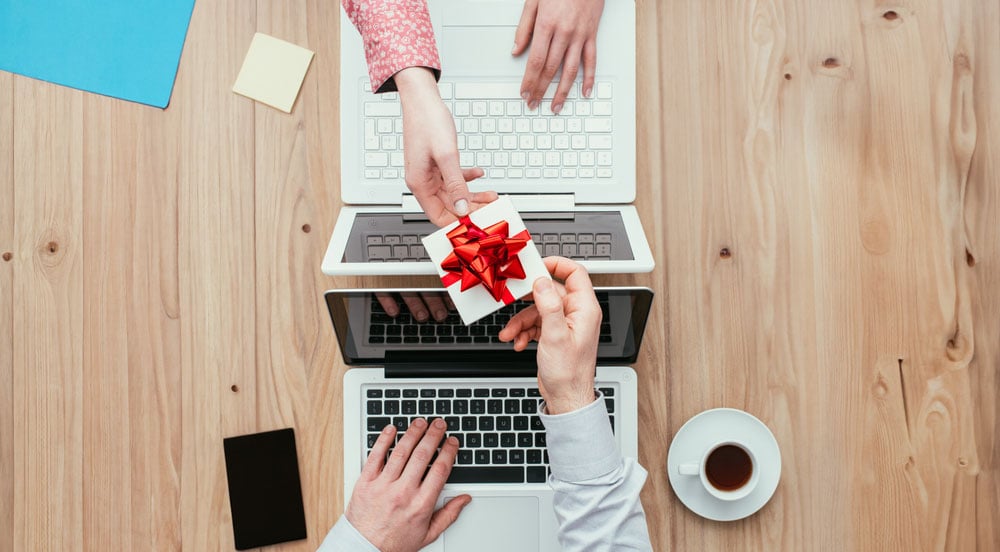 Foto tirada por cima de uma mesa com dois computadores e suas pessoas, sendo que uma está entregando o Vale-Presente Sem Parar Empresas para outra