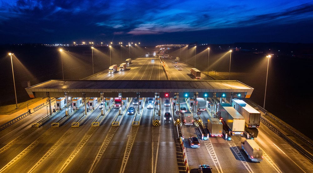 Praça de cobrança de rodovia à noite com carros e alguns caminhões passando na cabine de pagamento eletrônico, pois utilizam o vale-pedágio Sem Parar Empresas