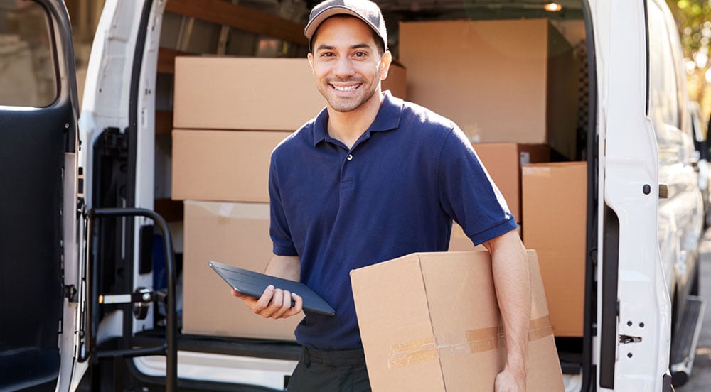 Homem segurando uma caixa e um tablet em frente a uma van carregada para fazer o serviço de entrega com a Sem Parar Empresas