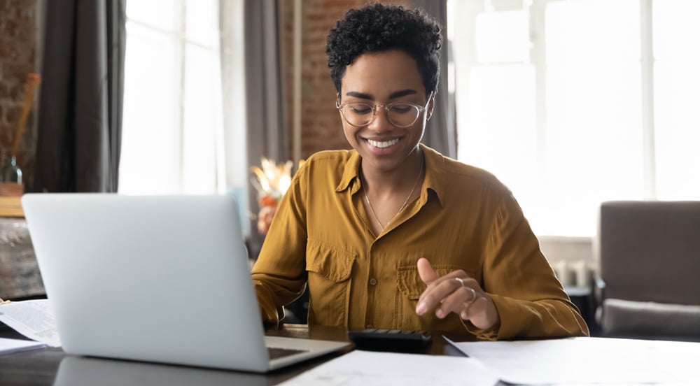 Mulher sorrindo em home office, atrás do seu notebook e usando uma calculadora para somar sua ajuda de custo.