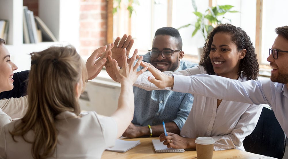 Sem Parar Empresas: cinco pessoas em mesa redonda dando as mãos no centro, pois concluíram seu programa de desenvolvimento profissional