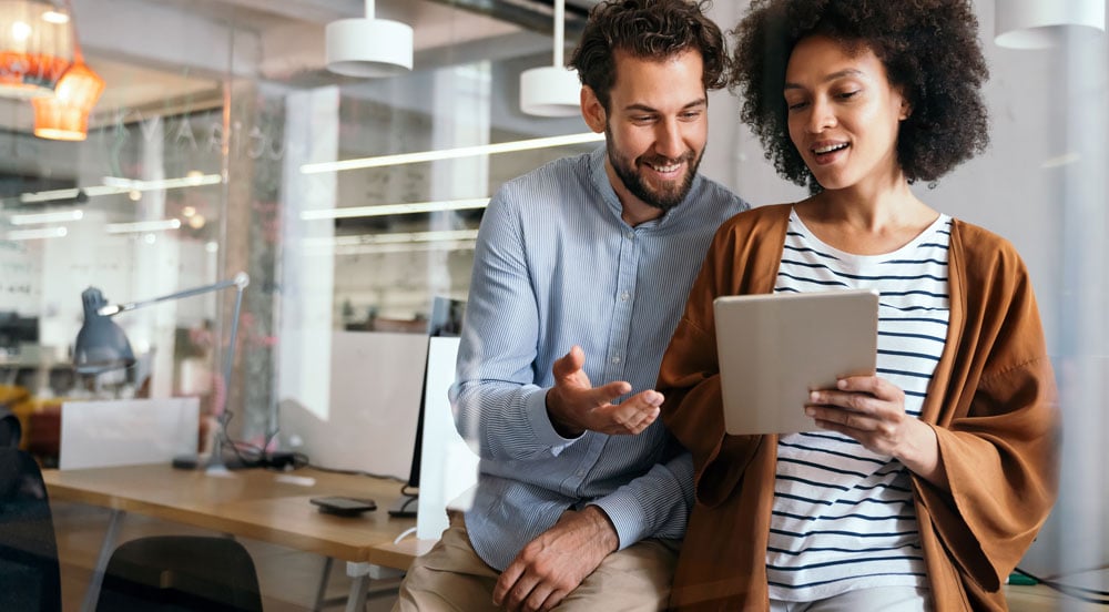Um homem e uma mulher em um escritório. Ela está segurando um tablet para analisar as soluções de benefícios da Sem Parar Empresas