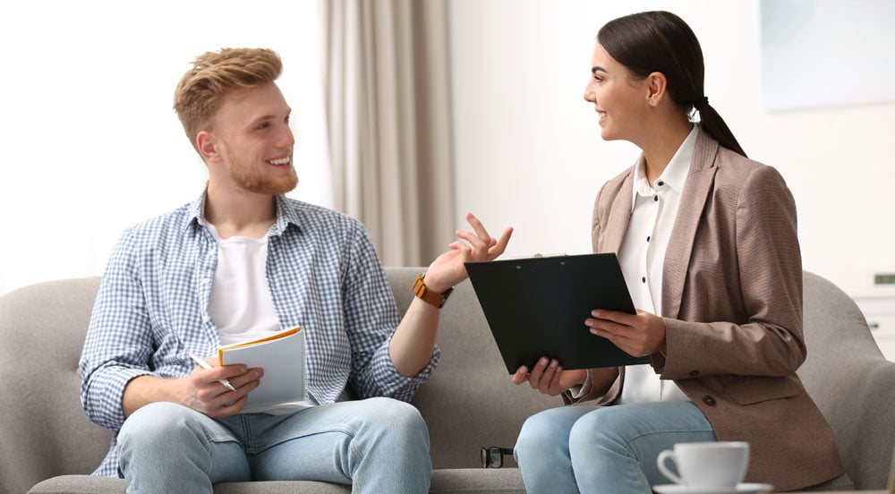 Homem e mulher que trabalham juntos sentados em um sofá, sorrindo e conversando sobre os cuidados com a saúde mental dos colaboradores
