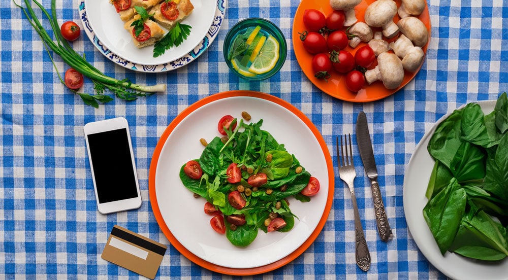 Mesa de um restaurante com uma toalha xadrez, pratos postos, talheres, um celular e um cartão refeição Sem Parar Empresas.