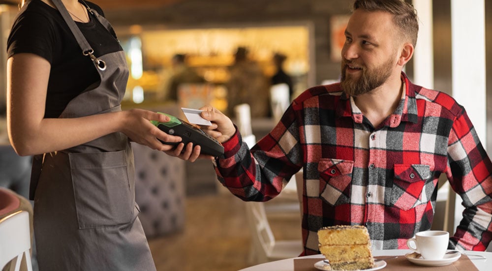 Um homem sentado à mesa em uma padaria fazendo o pagamento por cartão Flex Sem Parar Empresas para a garçonete