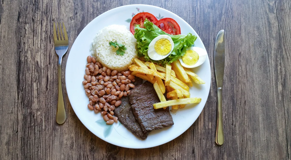 Sem Parar Empresas: garfo, faca e um prato com arroz, feijão, ovo, carne, batata frita e salada. A pessoa que vai comer está utilizando os benefícios do programa de alimentação do trabalhador.